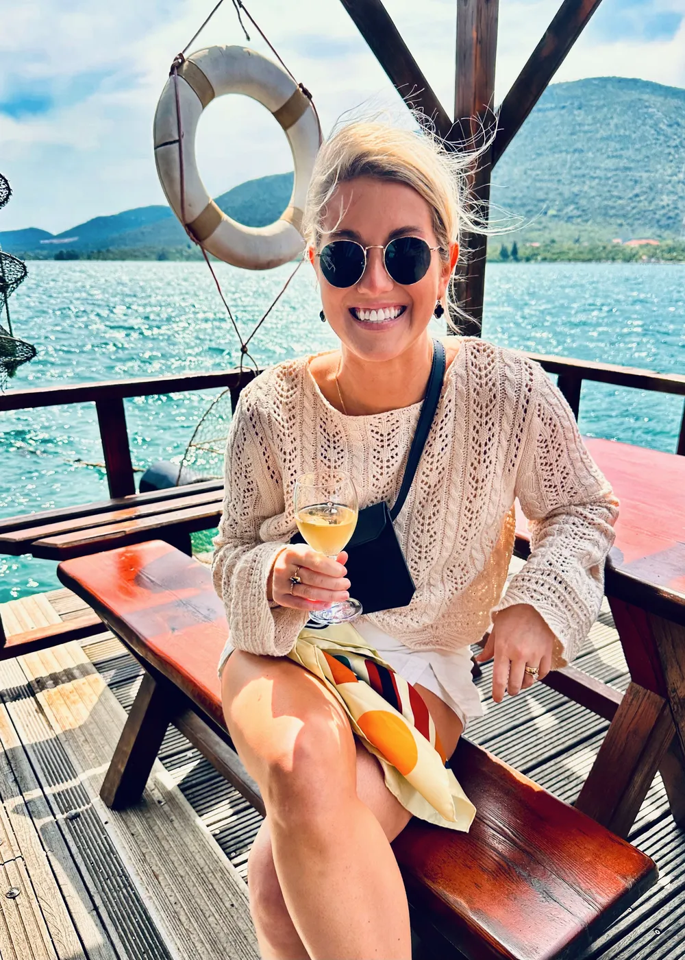 Kate Labourdette with a glass of wine on the deck of a boat, water and hills behind